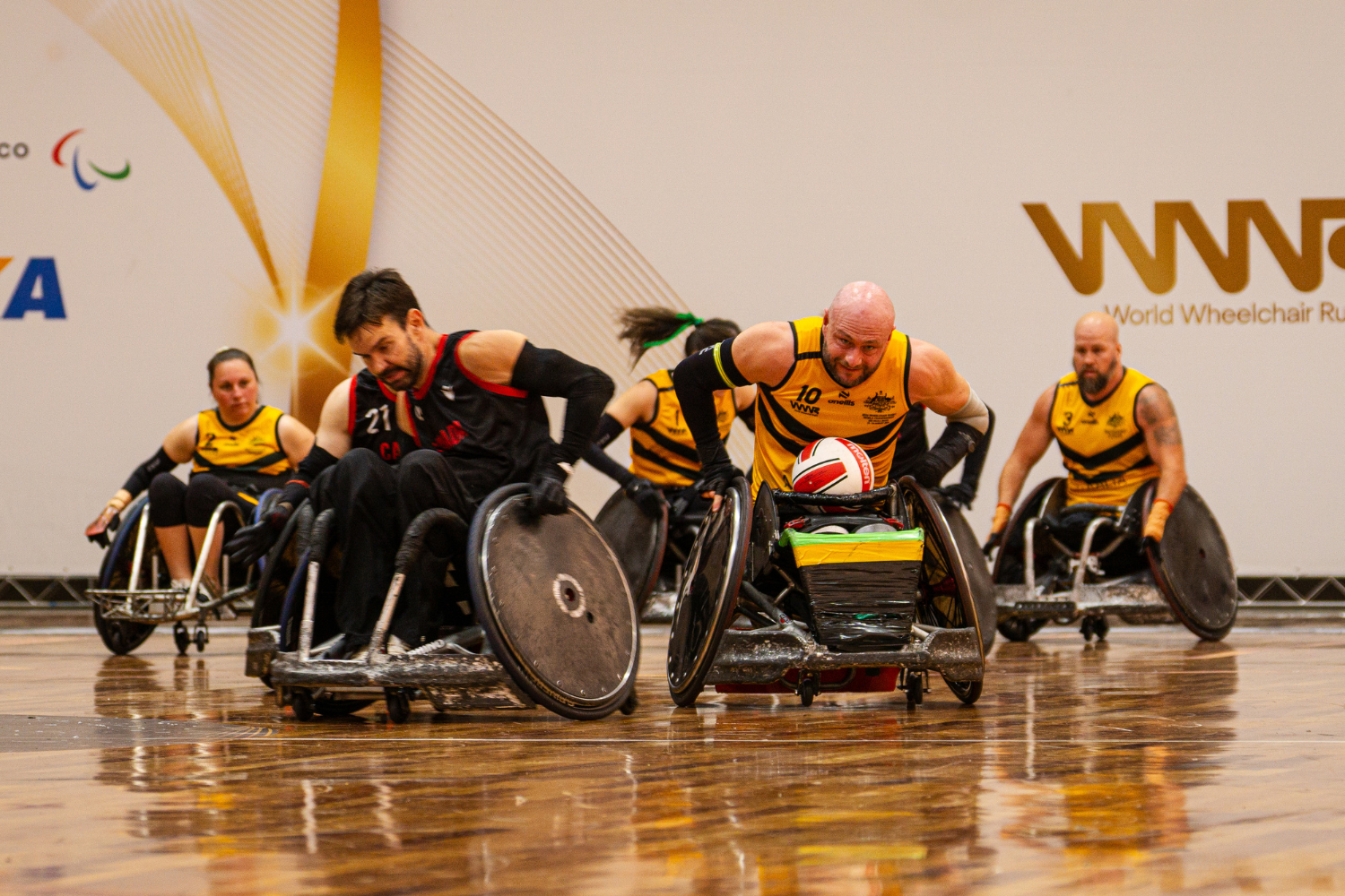Wheelchair rugby players race for the ball during a match on a wooden court, wearing yellow and black jerseys in an intense moment of action on wheels.