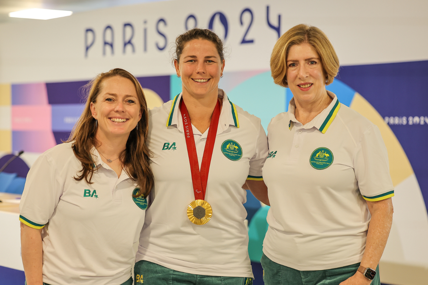 Three smiling athletes pose together; central athlete wears a gold medal at Paris 2024 backdrop.