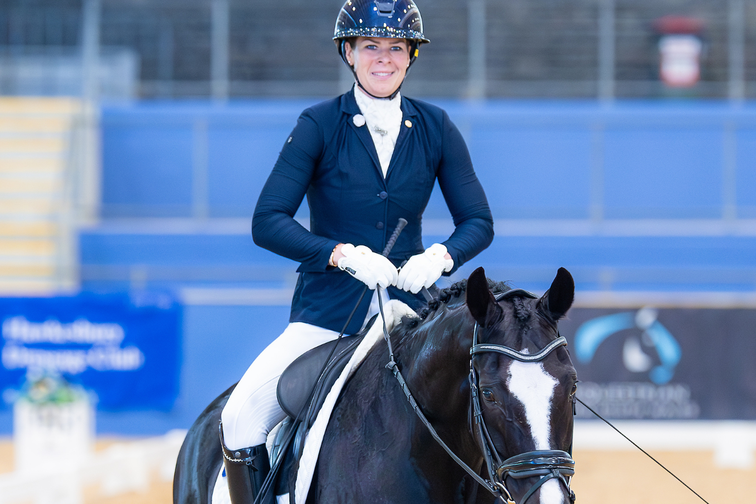 Female equestrian in a navy blazer and white gloves riding a black horse in an arena, smiling at the camera with a helmet on.