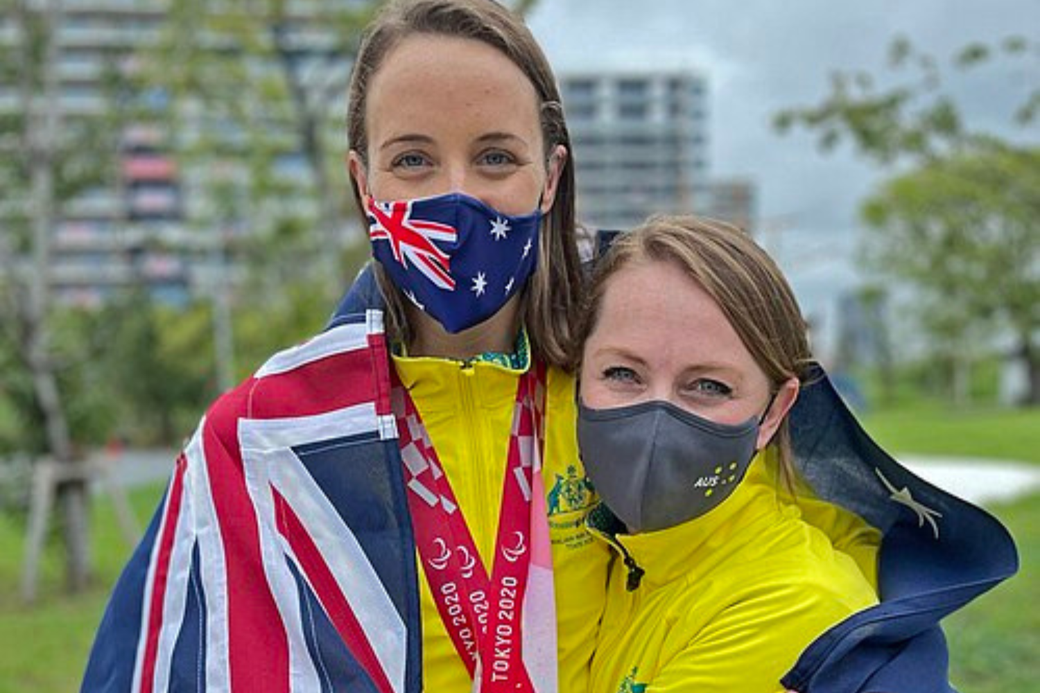 Two women outdoors in yellow jackets wear masks; one has an Australia flag mask and a Union Jack flag draped over her shoulders, with a Tokyo 2020 lanyard visible.