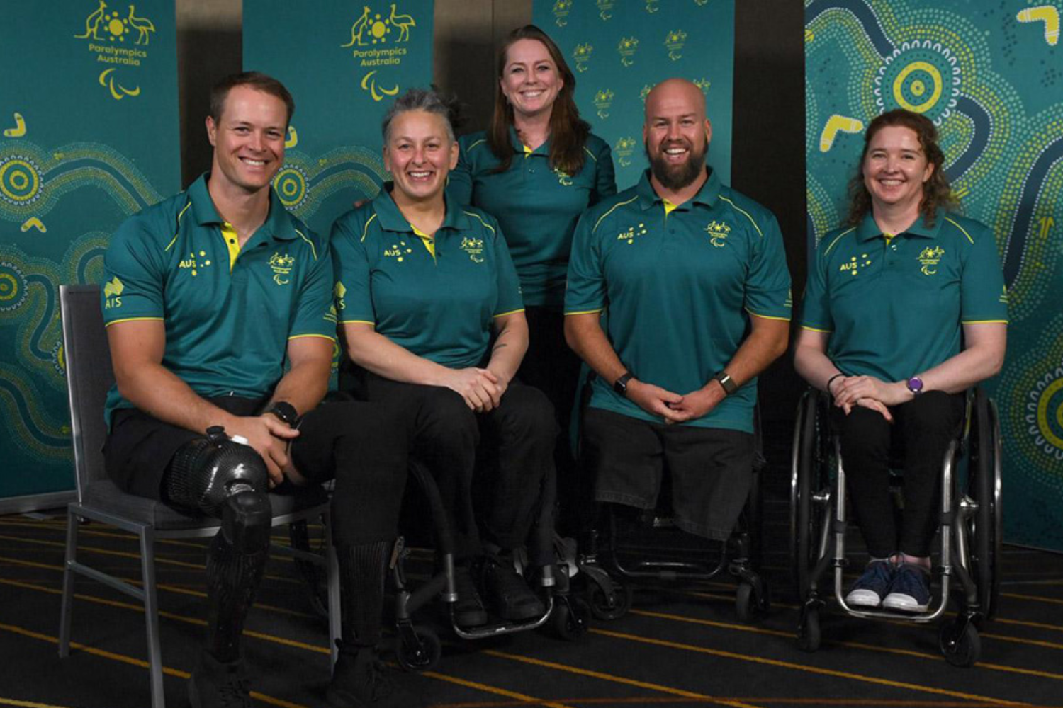 Group of five Paralympics Australia teammates posing for a photo, three seated on chairs and two in wheelchairs, wearing teal polo shirts with logos, in a teal patterned backdrop.