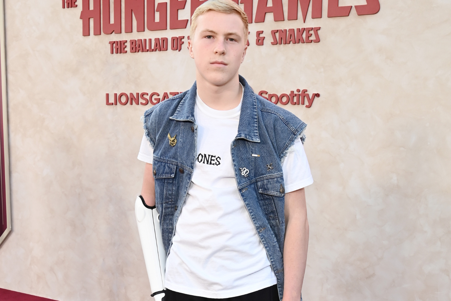 Young man in a white tee and sleeveless denim vest, carrying a white prosthetic arm brace, posing in front of a Hunger Games backdrop.