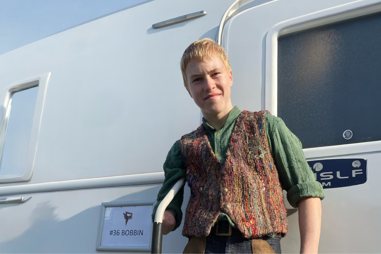 Young man in a colorful knitted vest leans against a white trailer, beside a sign that reads '#36 BOBBIN'.