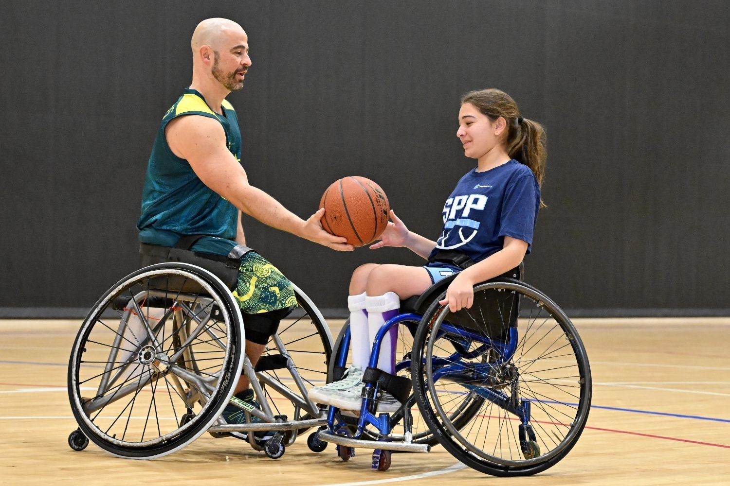 Two wheelchair basketball players on an indoor court exchange a basketball.