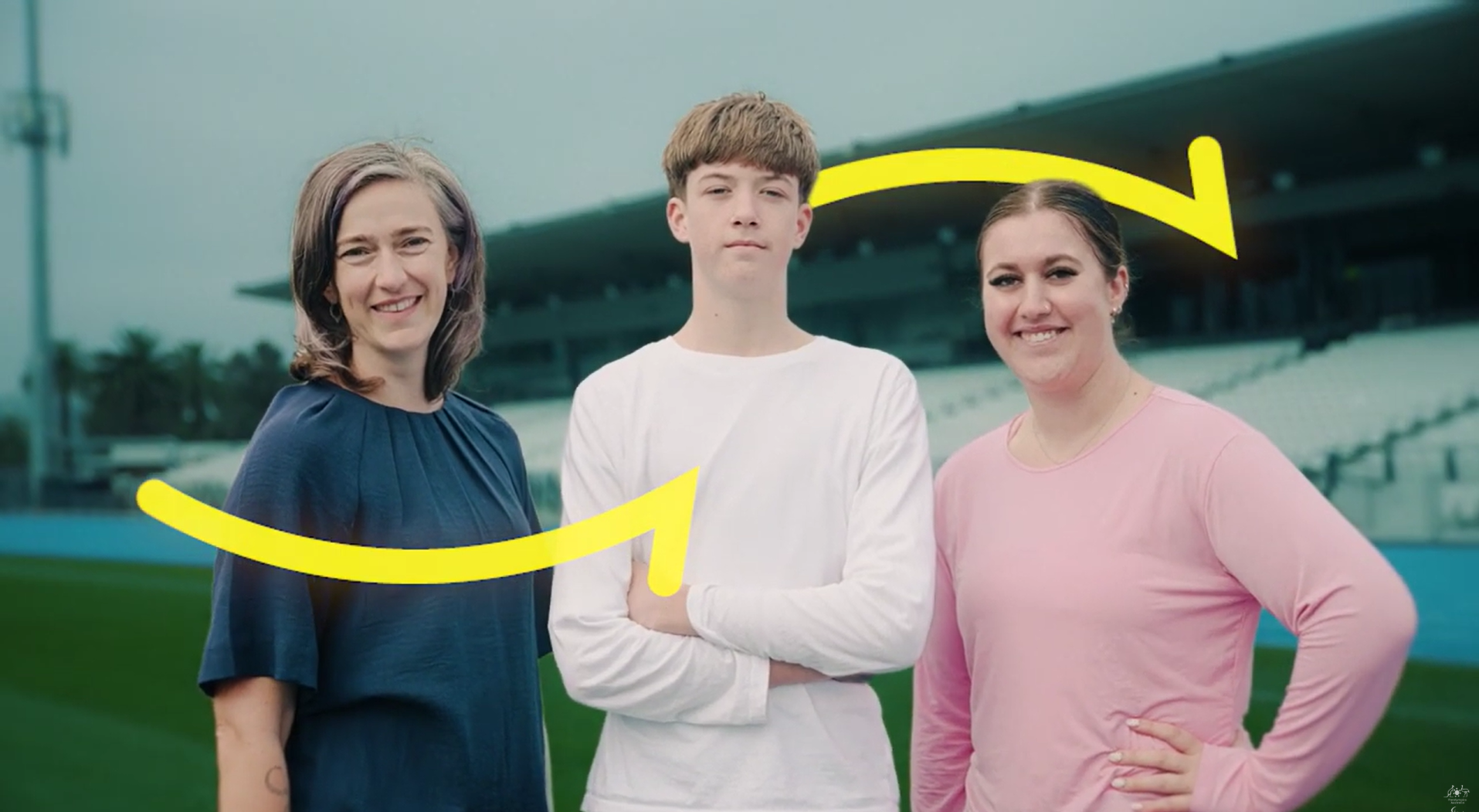 Three people pose on a football stadium field with two yellow curved arrows across the image.