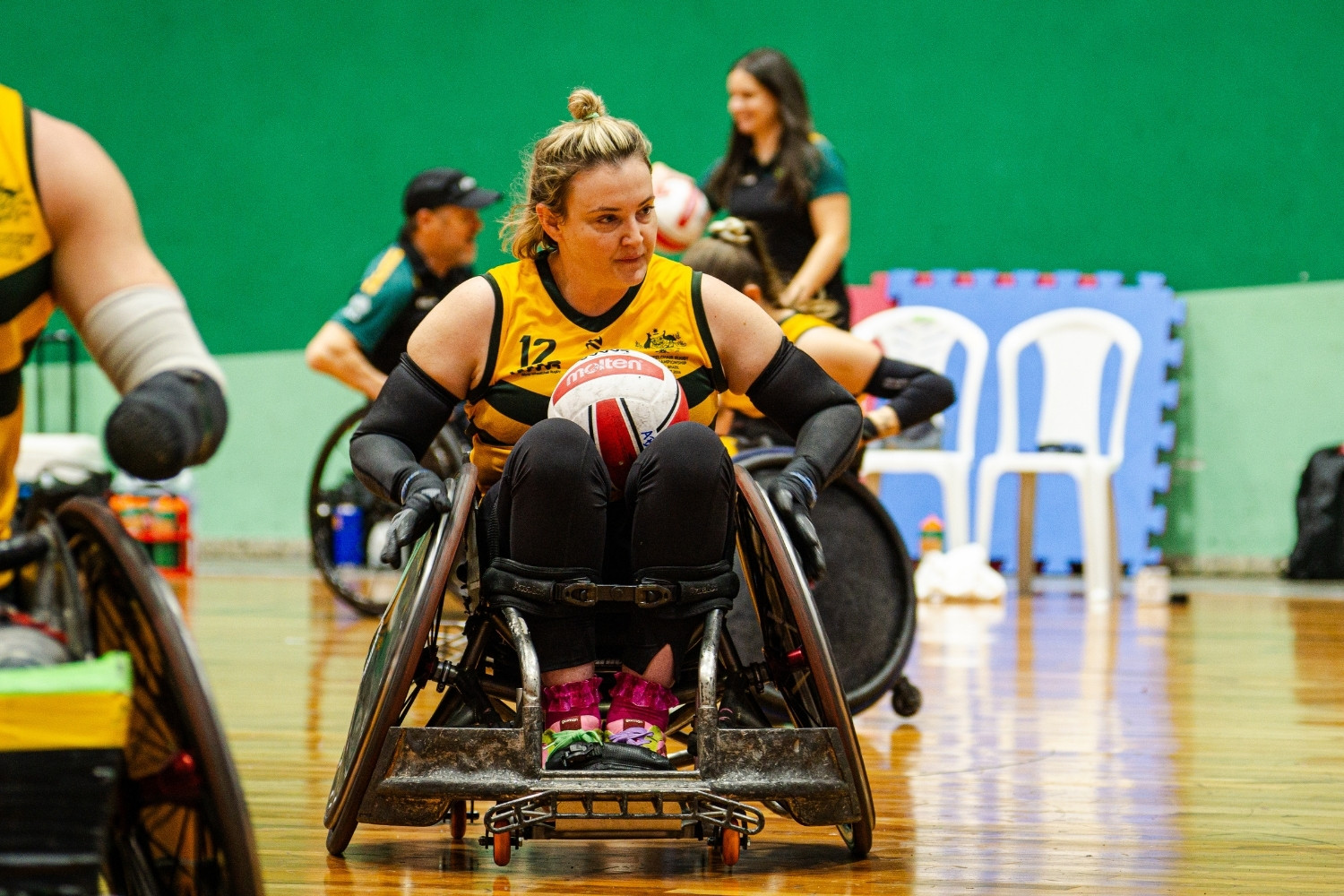 Female wheelchair rugby player in a yellow jersey grips a ball in her lap on an indoor court, focusing ahead as teammates watch in the background.
