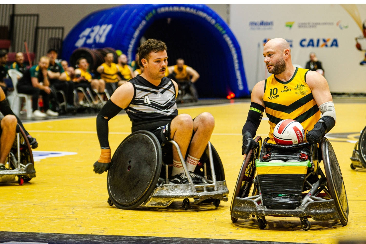 Wheelchair rugby players contest the ball on a bright yellow court; left player in black/gray with number 4, right player in yellow/green with number 10 holding the ball near the front stall.