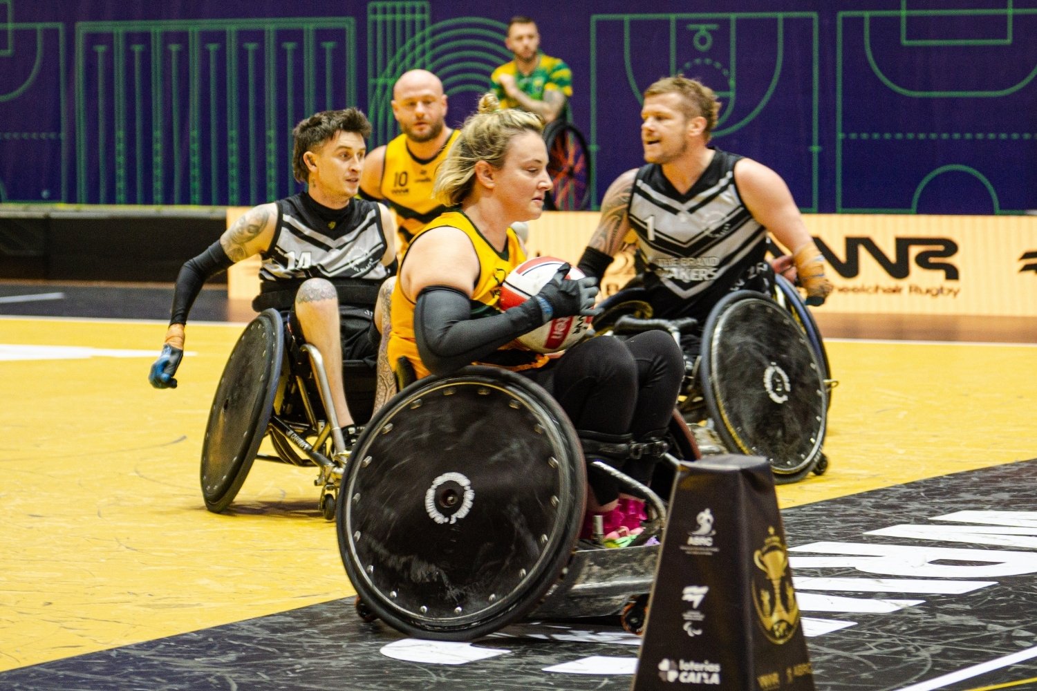 Female wheelchair rugby player in a yellow jersey advances with the ball while two opponents in black-and-white jerseys pursue on an indoor court.