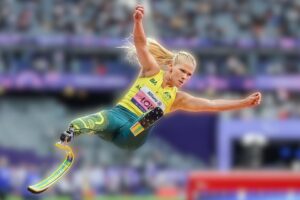 Female para-athlete in a yellow top and teal leggings soars mid-air for a long jump, powered by a curved prosthetic blade in a stadium setting.