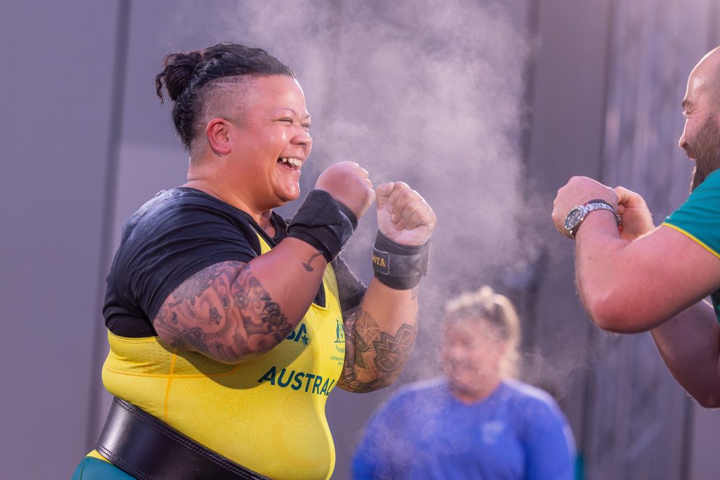 Athlete in a yellow singlet laughs and raises fists in a celebratory moment with a teammate nearby amid gym mist.