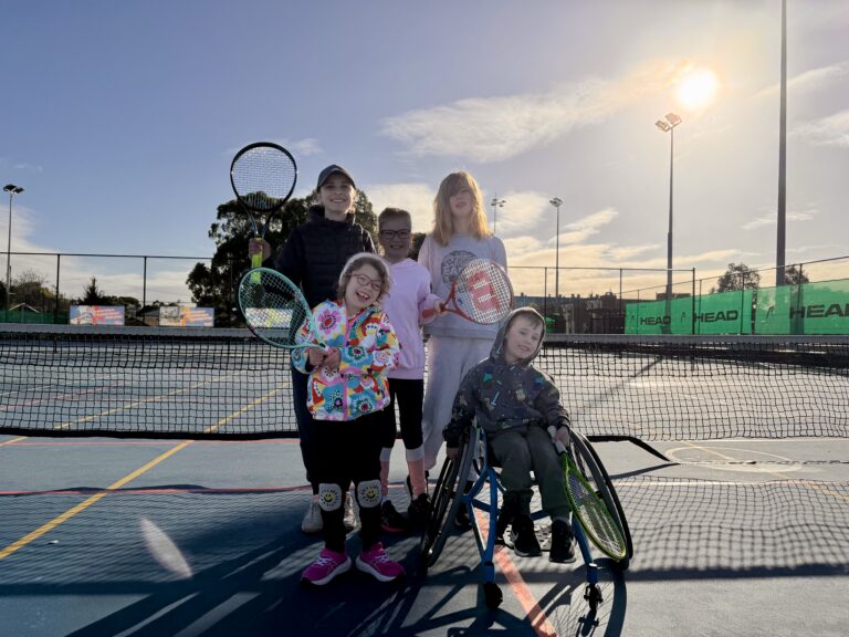 CCF166F5-1418-4808-9B1C-88BCF0698A90 | Paralympics Australia A group of kids on a tennis court posing with rackets, one in a wheelchair, sunny day and a chain-link fence in the background.