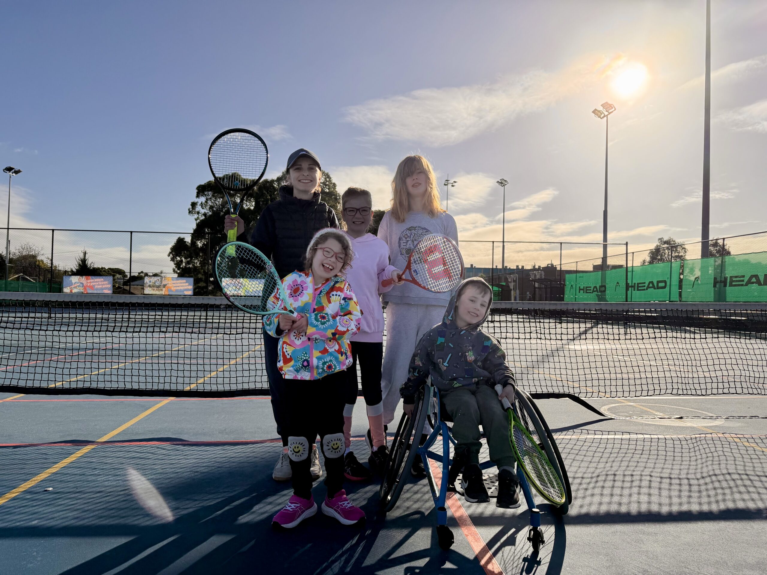 A group of kids on a tennis court posing with rackets, one in a wheelchair, sunny day and a chain-link fence in the background.