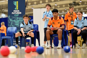 A young boccia player in a light blue polo, seated in a blue chair, watches his throw with his arm still extended at the Libby Kosmala Para Futures Cup. Athletes in light blue and orange polos look on, with red, blue and white boccia balls on the court in the foreground.
