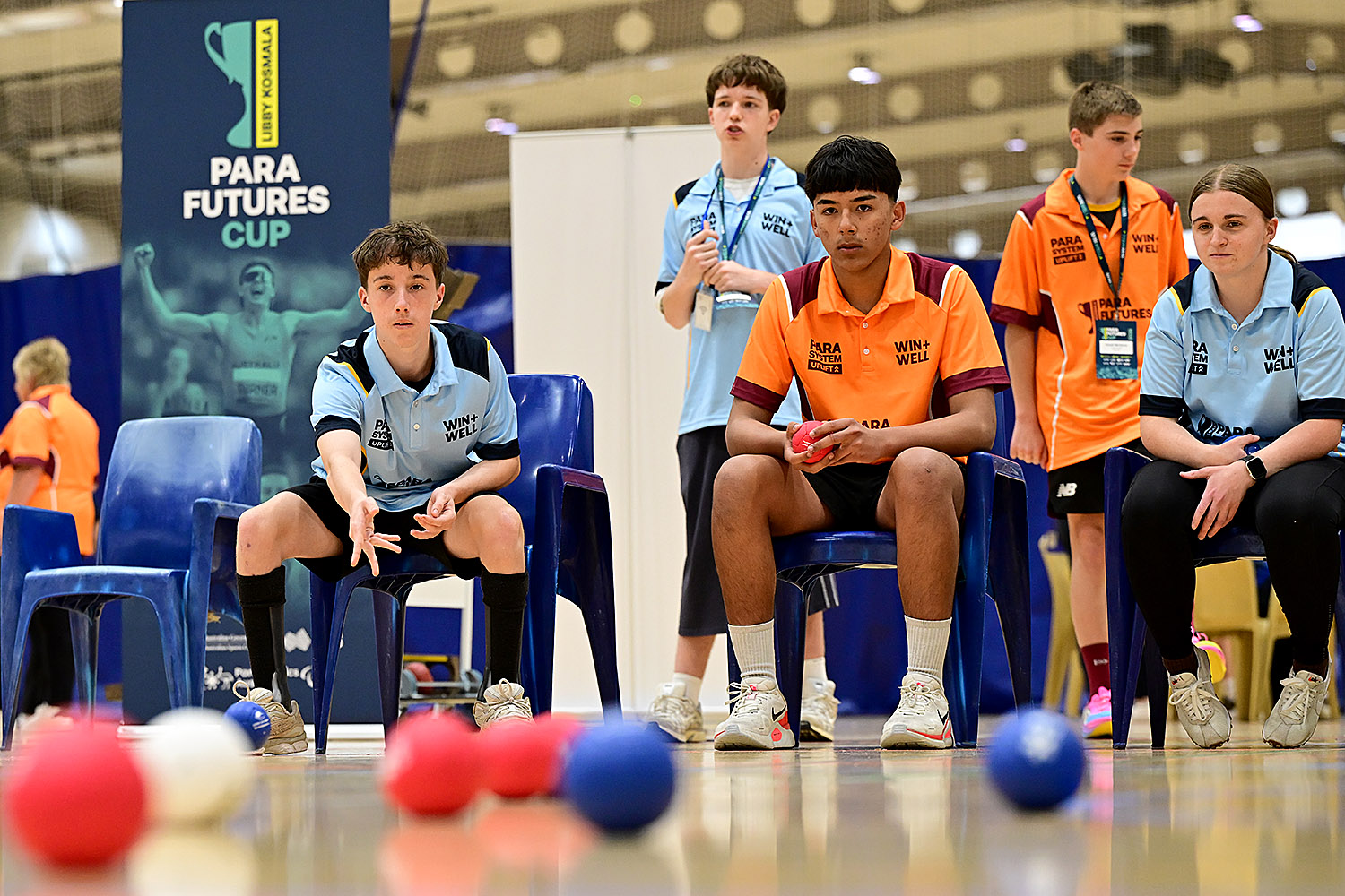 A young boccia player in a light blue polo, seated in a blue chair, watches his throw with his arm still extended at the Libby Kosmala Para Futures Cup. Athletes in light blue and orange polos look on, with red, blue and white boccia balls on the court in the foreground.